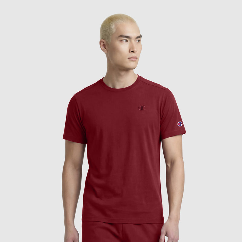 A person with short blond hair, wearing a maroon Champion Classic T-Shirt, C Logo, and matching maroon pants stands against a plain light gray background, looking slightly to the left.