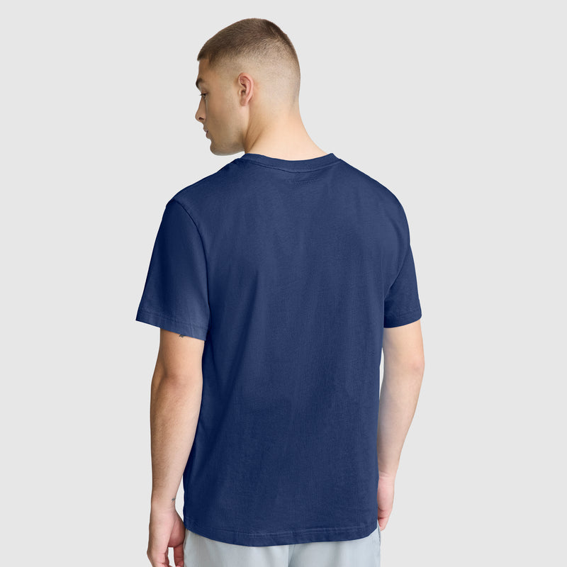 A man stands facing away, wearing the Champion Classic T-Shirt, C Logo, crafted from soft cotton fabric in plain navy blue, paired with light-colored pants against a light gray background.