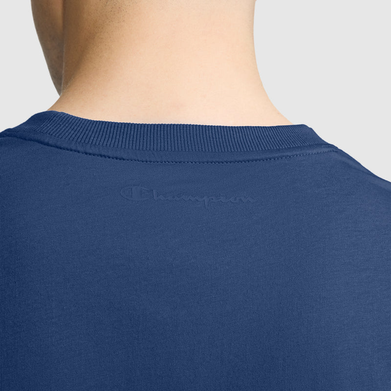 Close-up of the back of a person's neck and shoulders, wearing a blue Champion Classic T-Shirt, C Logo. The classic men's shirt features the Champion logo subtly embossed across the upper back. The background is plain light gray.