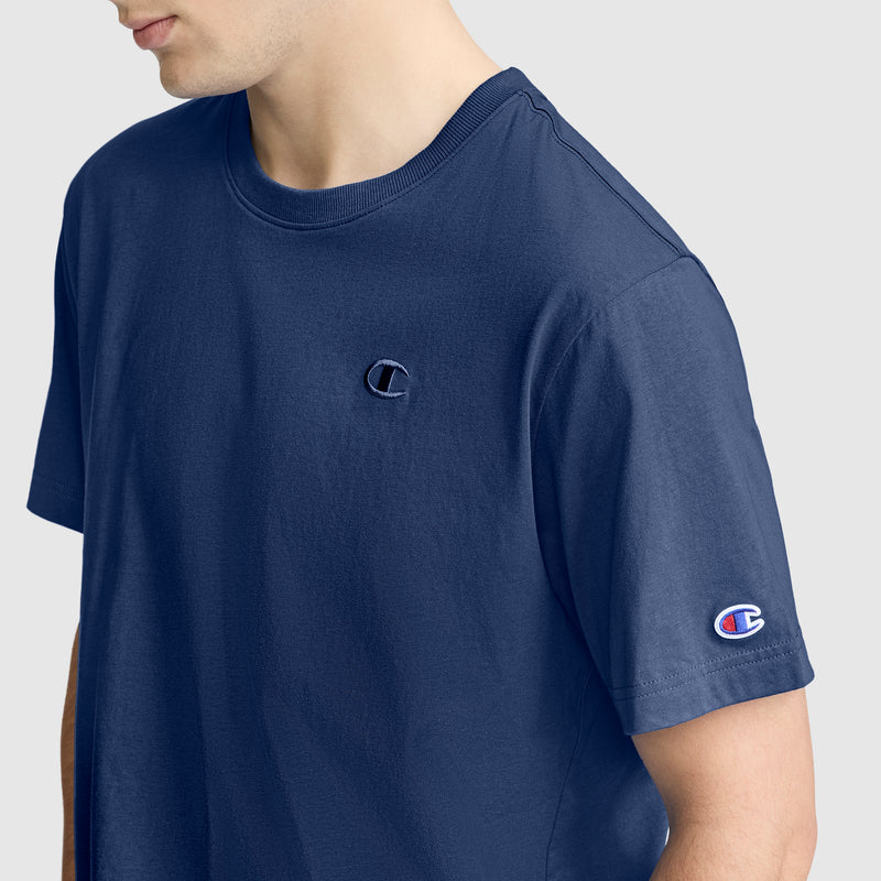 A person wearing a navy blue Classic T-Shirt, C Logo by Champion is shown from the shoulders to mid-torso, featuring small Champion logos on the chest and left sleeve. The background is plain light gray.