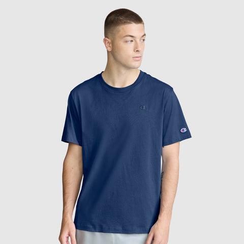 A young man wearing the Champion Classic T-Shirt, C Logo in navy blue—crafted from soft cotton with distinctive small Champion logos on the chest and sleeve—stands against a plain light gray background, looking to his left.