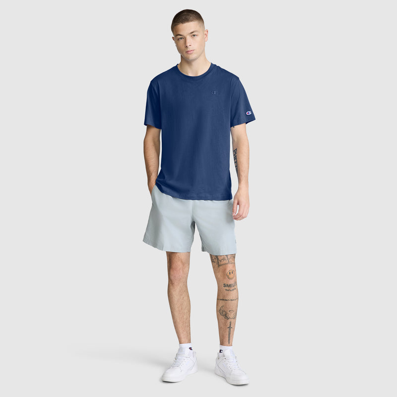 A young man with short hair stands facing forward against a plain light gray background, wearing the Champion Classic T-Shirt, C Logo in navy blue paired with light gray shorts and white sneakers. He has one hand in his pocket and tattoos visible on his left leg and arm.