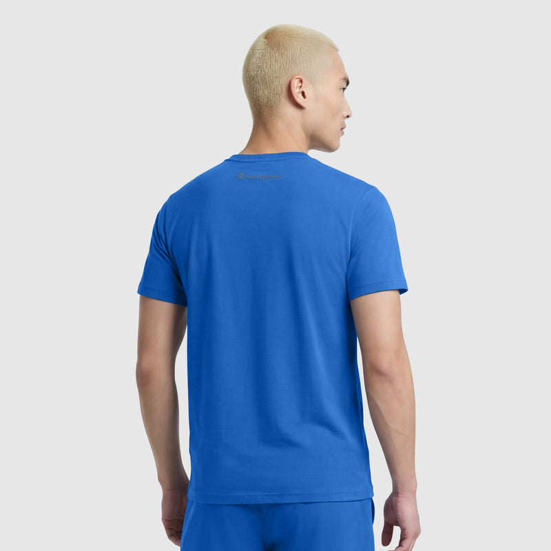 A person with short, bleached blonde hair faces away from the camera, wearing a bright blue Champion Classic T-Shirt, C Logo, with matching blue shorts against a plain light background.