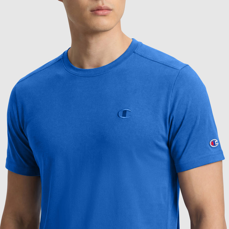 A man wears a bright blue Champion Classic T-Shirt, C Logo, with the brand’s chest and sleeve logos visible, shown from the chest up against a light gray background.