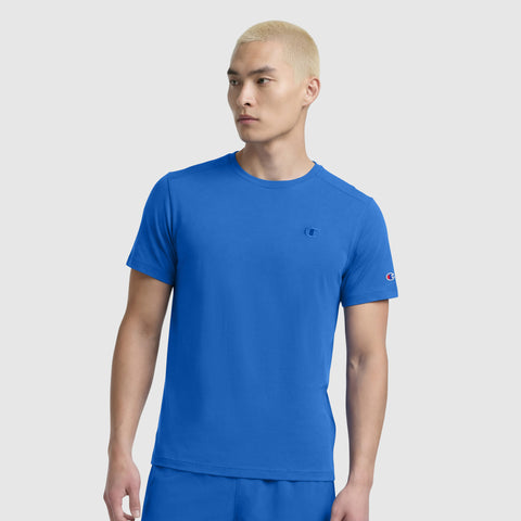 A person with short, bleached blond hair wears a bright blue Champion Classic T-Shirt with a C Logo, paired with matching shorts, standing against a plain light gray background and looking slightly to the side.