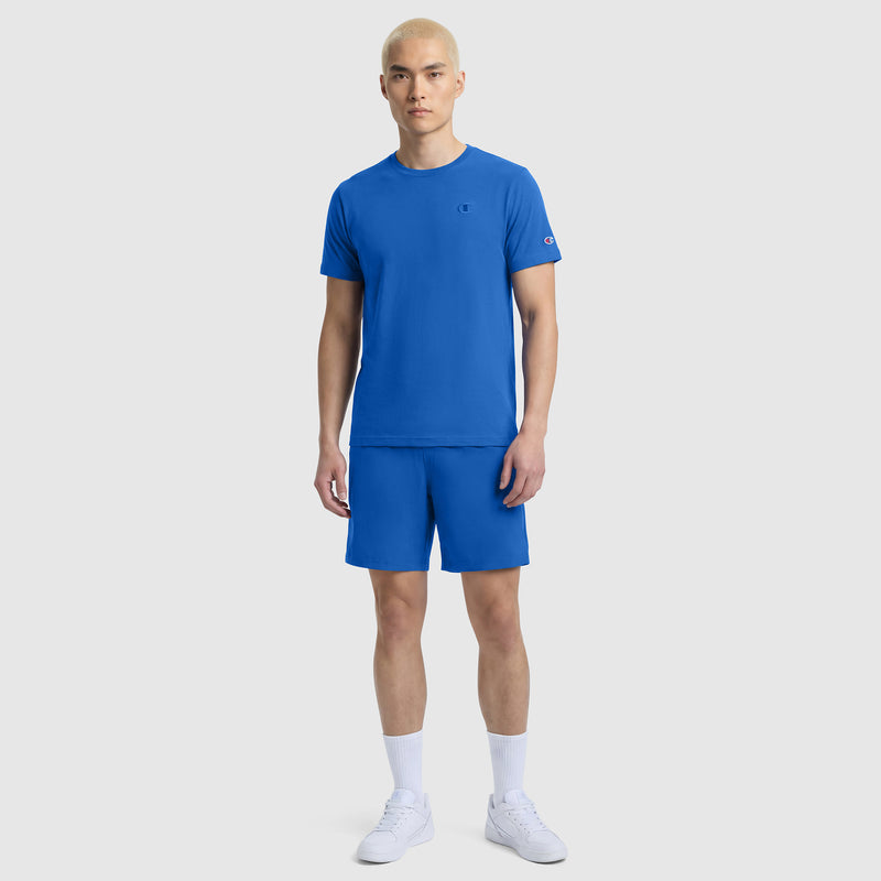 A person with short blond hair wears the Classic T-Shirt, C Logo by Champion with matching blue shorts, white socks, and sneakers, standing against a plain light gray background.