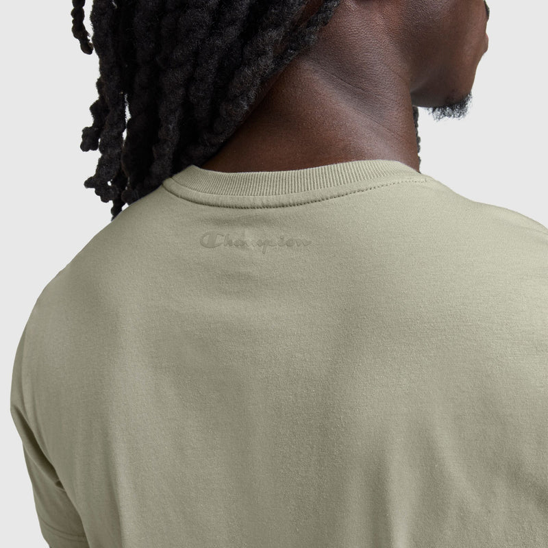 A close-up of a person with dreadlocks wearing the Champion Classic T-Shirt, C Logo in light beige cotton. “Champion” is subtly embossed near the neckline, giving this classic men's shirt a refined touch against a plain, light backdrop.