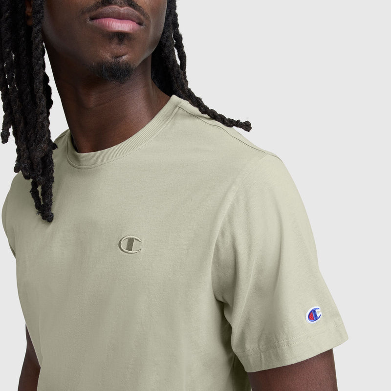 A person with long, braided hair wears the Champion Classic T-Shirt, C Logo in light beige, featuring the embroidered Champion C logo on the chest and sleeve. Their face is partially visible.