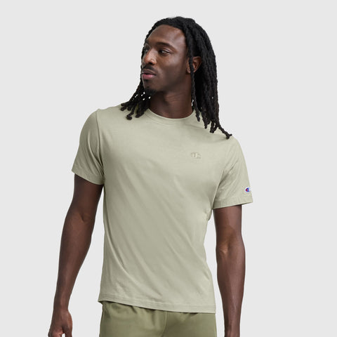 A man with long dreadlocks stands against a plain light gray background, wearing the Champion Classic T-Shirt, C Logo, and matching shorts, looking off to the side.