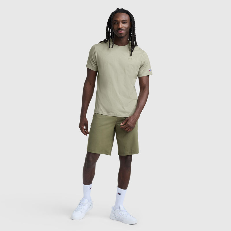 A man with long braids wears a light olive green Classic T-Shirt by Champion (C Logo), olive green shorts, white crew socks, and white sneakers. He stands on a plain white background, smiling with one hand in his pocket.