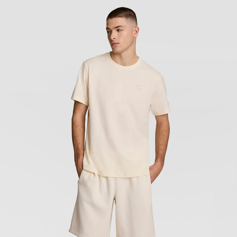 A young man stands with his hands in his shorts pockets, wearing a Champion Classic T-Shirt with the C Logo and matching light beige shorts against a plain light gray background.