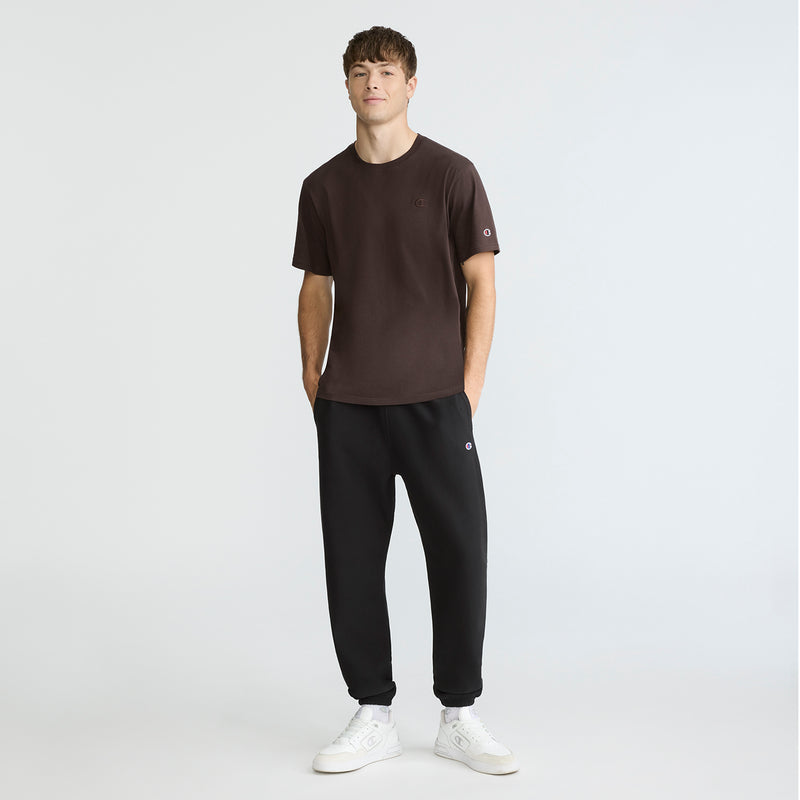 A young man stands against a plain background, wearing a dark brown Champion Classic T-Shirt, C Logo, paired with black jogger pants and white sneakers. His hands are in his pockets as he looks ahead with a neutral expression.