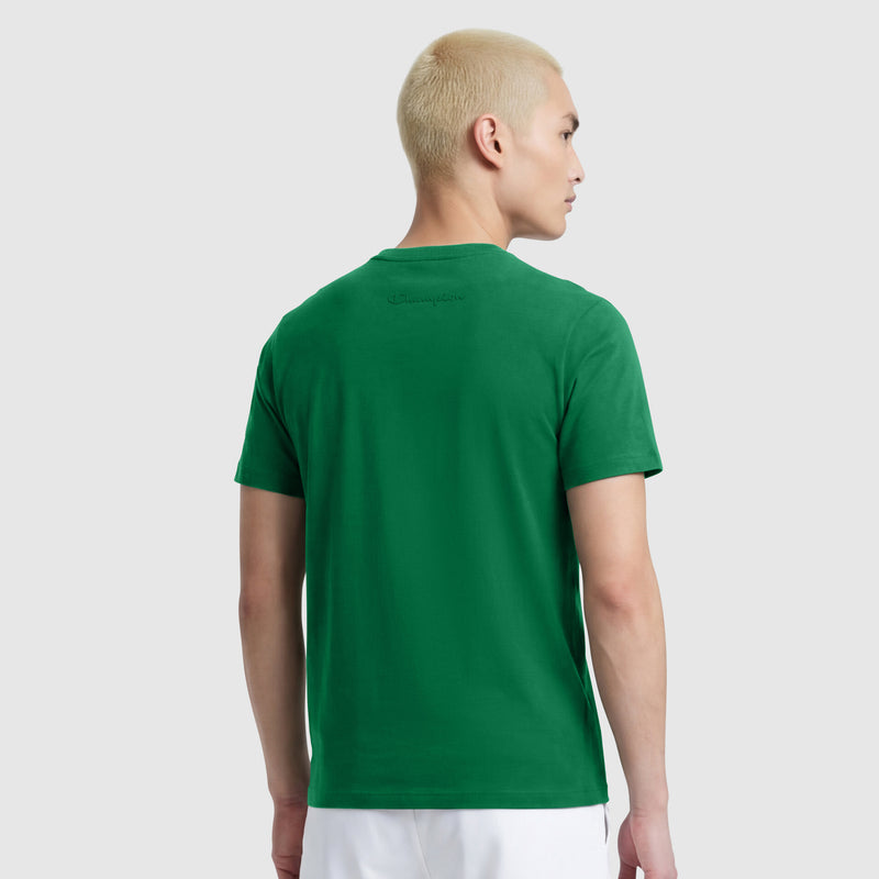 A person with short, bleached blonde hair faces away, wearing a Champion Classic T-Shirt, C Logo in green with a crewneck, paired with white pants against a light gray background.