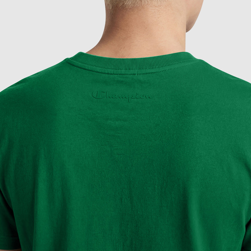 A person is seen from behind wearing a green Champion Classic T-Shirt, C Logo. "Champion" is embroidered in green thread just below the collar on the upper back of this soft cotton tee.