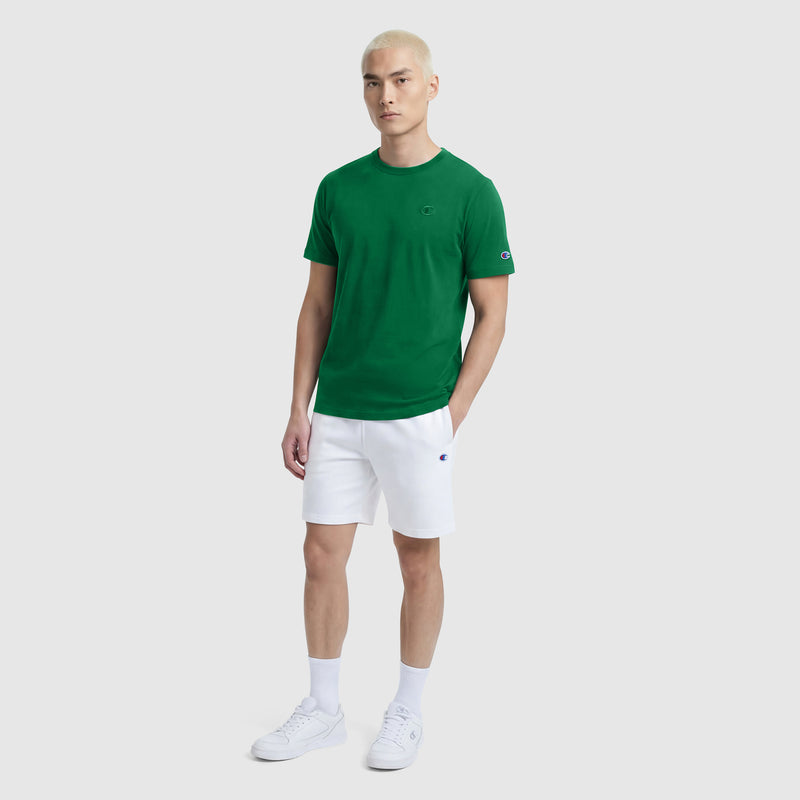 A person with short blonde hair wears a Champion Classic T-Shirt with the C Logo in green, paired with white shorts, socks, and sneakers, standing against a plain light gray background.