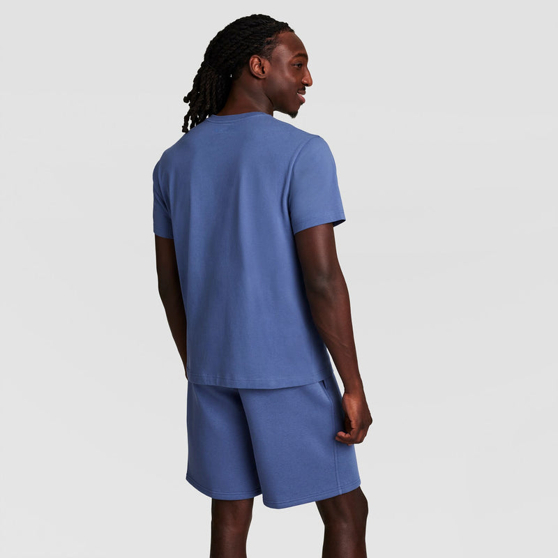 A man with long braided hair faces away, wearing a matching blue Champion Classic T-Shirt with C Logo and shorts set, against a plain light background.