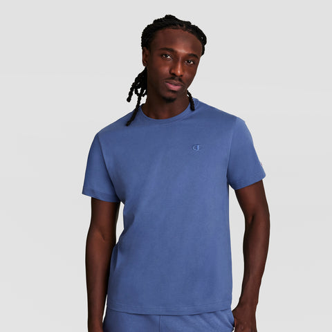 A man with long braided hair is wearing a Champion Classic T-Shirt, C Logo, paired with matching blue pants, standing against a plain light gray background.