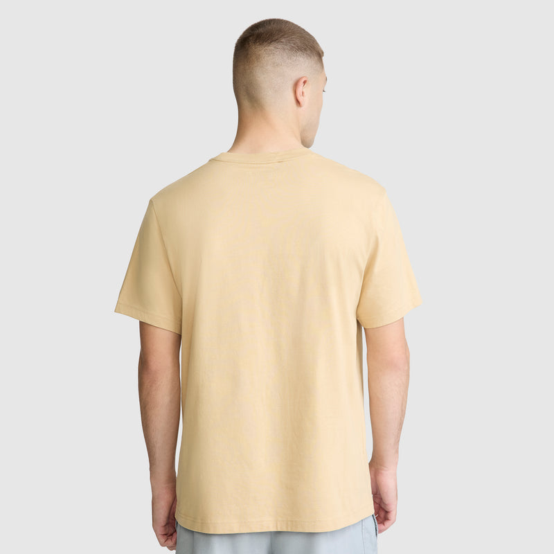 A person with short hair is standing with their back to the camera, wearing a beige Champion Classic T-Shirt, C Logo, and light-colored shorts against a plain gray background.