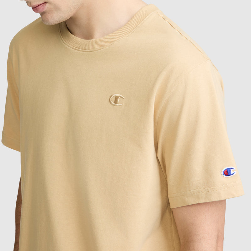 A person wearing the Champion Classic T-Shirt, C Logo in beige, displaying the embroidered Champion logo on both the chest and sleeve against a plain light background.