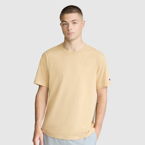A young man with short hair wears the Champion Classic T-Shirt featuring the C Logo in beige, paired with light pants. He stands against a plain gray background, looking at the camera with a neutral expression and one hand in his pocket.