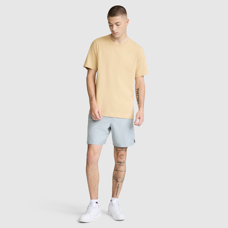 A young man stands against a plain background wearing the Champion Classic T-Shirt, C Logo in beige, paired with light blue shorts and white sneakers. Tattoos are visible on his right arm and both legs as he looks slightly downward with one hand by his side.