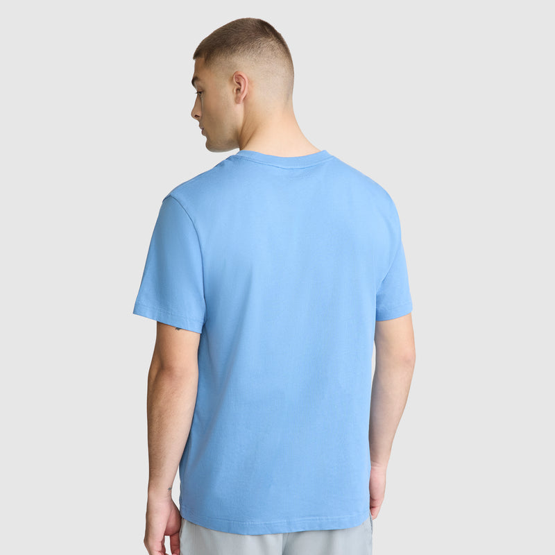 A man with short hair, facing away, is wearing the Champion Classic T-Shirt, C Logo in light blue and light-colored pants against a plain light gray background.