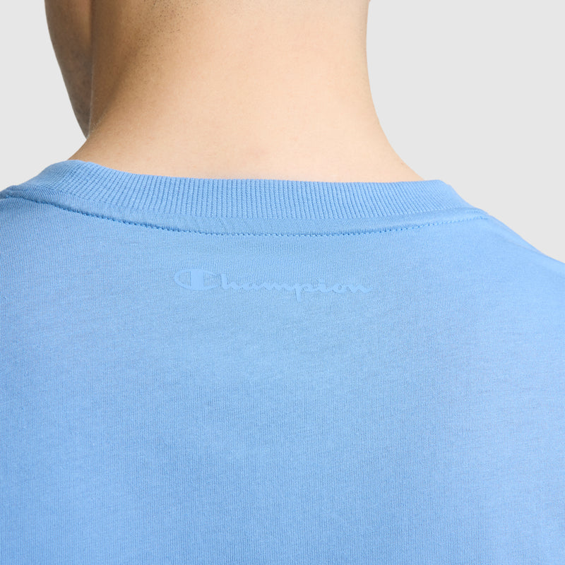 Close-up of the back of a person’s neck and shoulders wearing the Champion Classic T-Shirt, C Logo in light blue, featuring the faintly printed Champion branding near the collar.