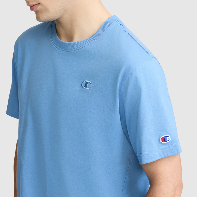 A person is wearing a light blue Champion Classic T-Shirt, C Logo for men. The Champion logo appears on the left chest and the left sleeve. This classic men's tee from Champion is crafted from soft cotton, and the background is plain and light-colored.