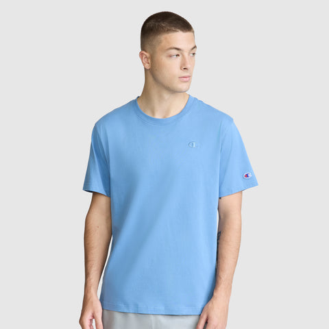 A young man wearing a light blue Champion Classic T-Shirt, C Logo, and light gray pants stands against a plain white background, looking to his left.