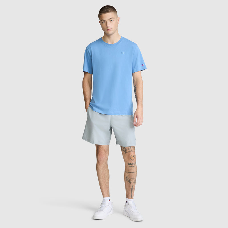 A young man stands facing forward with one hand in his pocket, wearing the Champion Classic T-Shirt, C Logo in light blue, paired with light gray shorts and white sneakers. He has tattoos on his legs and a neutral expression.