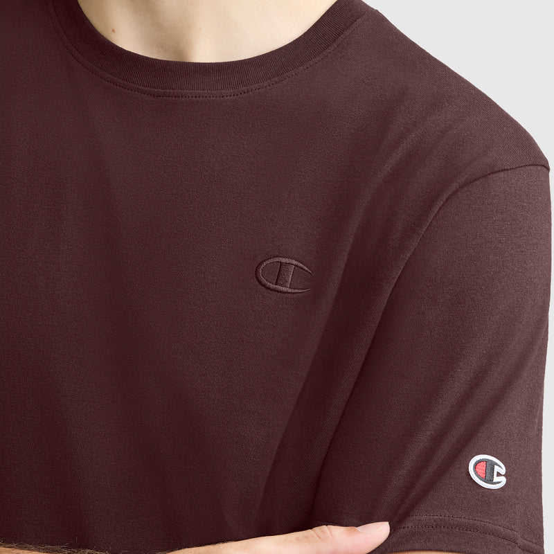 A person wearing the Champion Classic T-Shirt, C Logo in maroon, featuring an embroidered Champion logo on the chest and a small logo patch on the sleeve. This classic men's tee combines comfort with iconic style.