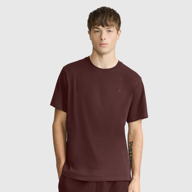 A young man with short brown hair wears a Champion Classic T-Shirt with C Logo in maroon, paired with matching pants, as he stands against a light gray background looking directly at the camera with a neutral expression.