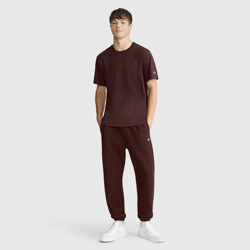 A person stands against a plain background wearing the Champion Classic T-Shirt, C Logo, paired with matching dark maroon sweatpants and white sneakers. Crafted from soft cotton fabric for everyday comfort, the look features a classic style with hands in pockets as the individual faces forward with a neutral expression.