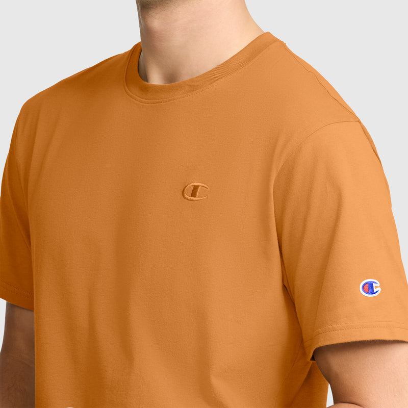 A person is wearing an orange Classic T-Shirt, C Logo by Champion, visible from shoulders to chest. The Champion logo is embroidered on the chest and the sleeve of this cotton T-shirt. The background is plain and light gray.
