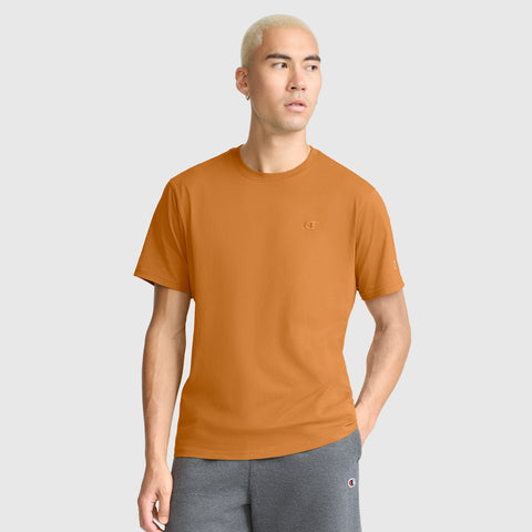 A man with light blond hair stands against a plain light background, wearing a Champion Classic T-Shirt, C Logo in orange paired with gray shorts. He has one hand in his pocket and gazes slightly to the side. The soft cotton fabric of the T-shirt ensures all-day comfort, adding to his effortlessly relaxed appearance.