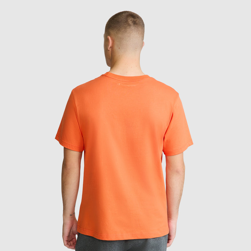 A person with short hair is standing facing away, wearing a Champion Classic T-Shirt, C Logo in orange made from soft cotton and dark pants against a plain light gray background.