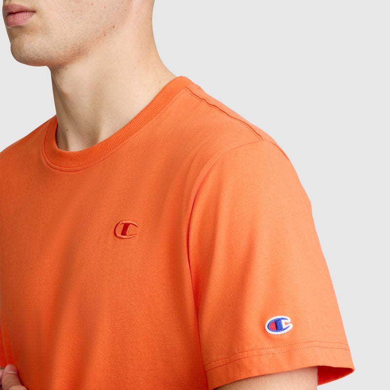 A person is wearing the Champion Classic T-Shirt, C Logo in orange, crafted from soft cotton fabric, featuring the signature C logo on the chest and sleeve. The background is a plain light gray.