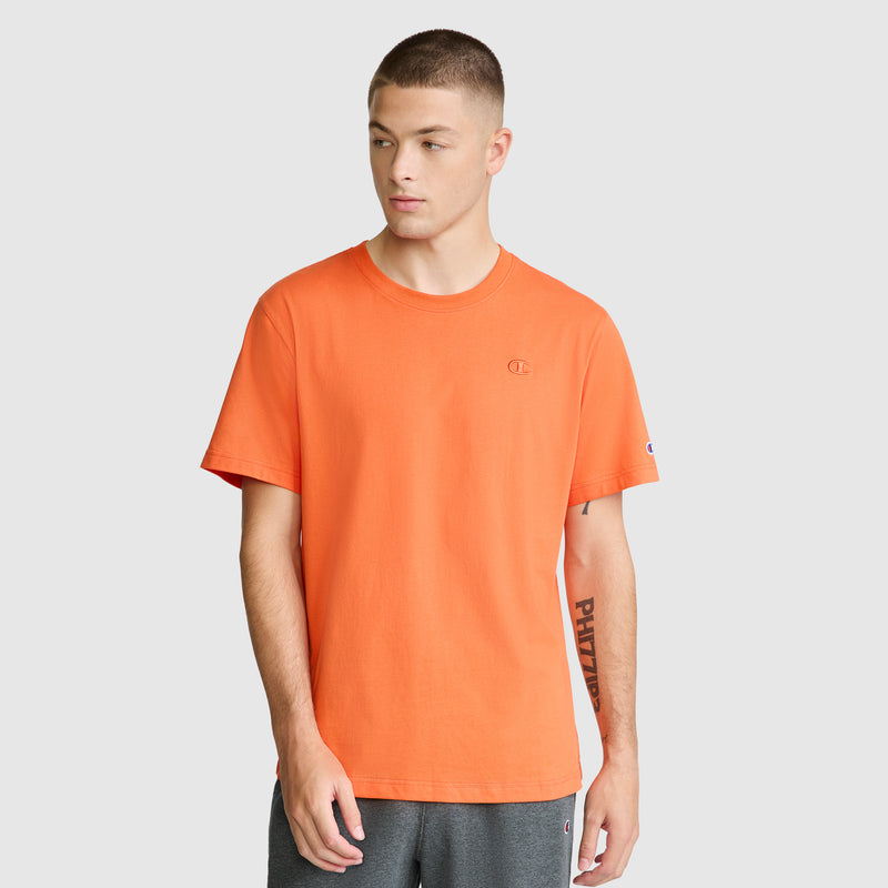 A young man wearing a Champion Classic T-Shirt, C Logo in orange and gray shorts stands against a plain light background, looking to his left with his hands by his sides. The soft cotton fabric of the Champion Classic T-Shirt ensures comfort while complementing his effortlessly cool style.