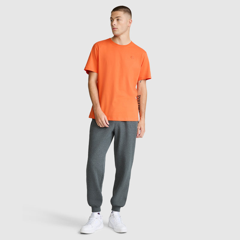 A person stands against a plain background wearing the Champion Classic T-Shirt, C Logo in orange, made from soft cotton fabric. They’ve paired it with gray jogger pants and white sneakers, looking off to the side with one hand by their side and the other resting near their pocket.