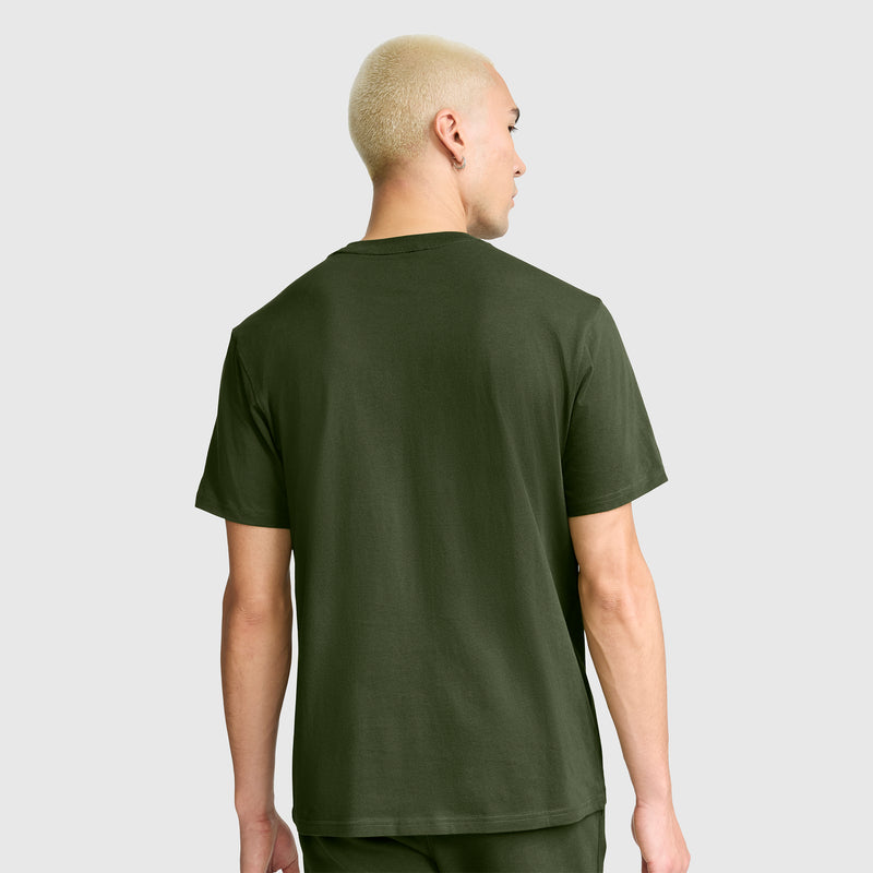 A person with short, bleached blonde hair is shown from the back, wearing a Champion Classic T-Shirt with C Logo in olive green and matching pants against a plain light gray background.