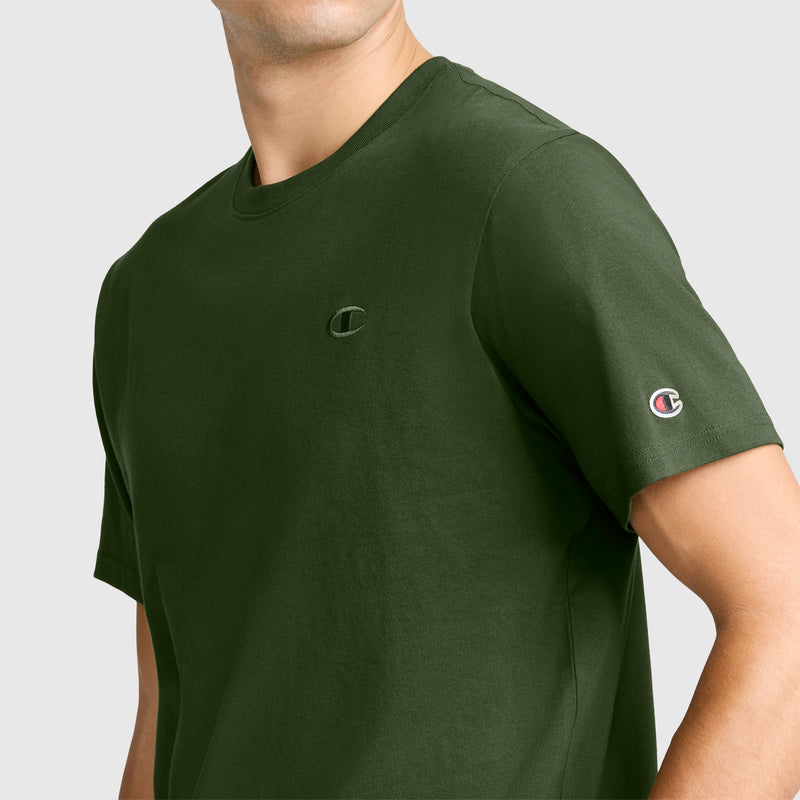A man wears the Champion Classic T-Shirt, C Logo in dark green, featuring the iconic Champion branding on the chest and left sleeve. The shirt is set against a plain light gray background and is crafted from soft cotton for all-day comfort.