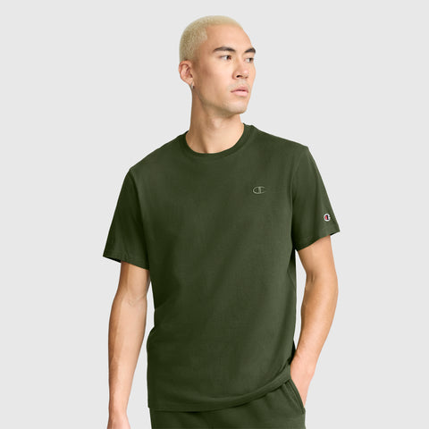 A person with short blond hair wears a Champion Classic T-Shirt featuring the C Logo in olive green cotton along with matching pants, standing with one hand in their pocket and gazing to the side against a plain background.