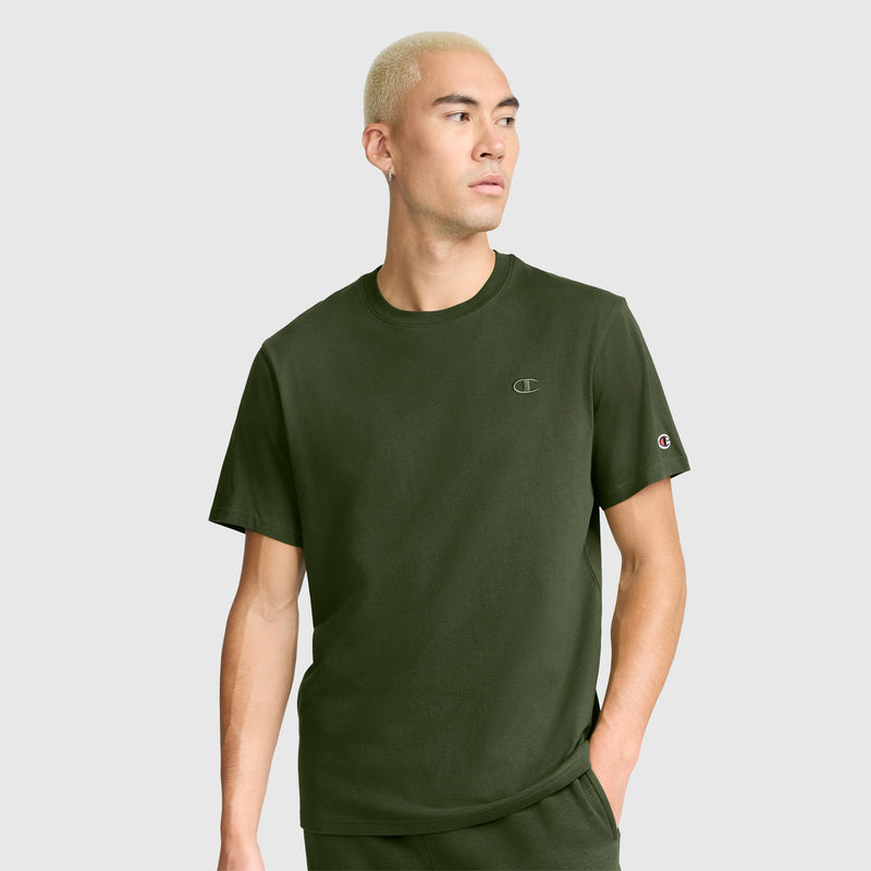 A person with short blond hair wears a Champion Classic T-Shirt featuring the C Logo in olive green cotton along with matching pants, standing with one hand in their pocket and gazing to the side against a plain background.