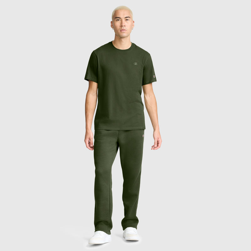 A person with short blond hair wears a dark green Champion Classic T-Shirt featuring the C Logo, paired with matching pants and white sneakers, standing against a plain light gray background.