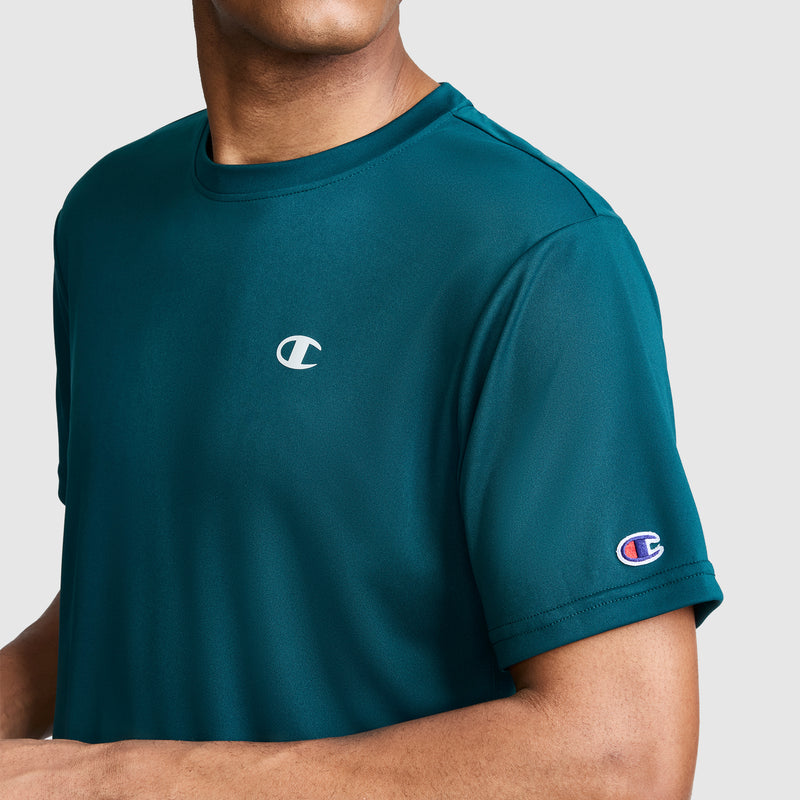 A person wearing a teal Champion Sport T-Shirt, Reflective C Logo—a men’s athletic shirt featuring embroidered logos on the chest and sleeve—stands out against a plain light gray background.