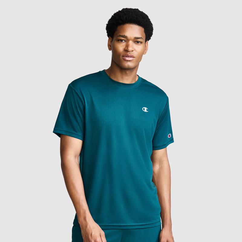 A man wearing a teal Champion Sport T-Shirt with a reflective C logo stands against a plain light gray background, looking at the camera with a neutral expression.