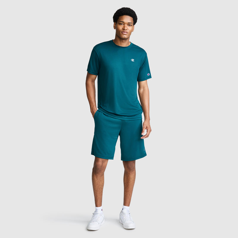 A man stands against a light gray background wearing the Champion Sport T-Shirt with Reflective C Logo in teal, paired with matching athletic shorts and white sneakers. The Sport T-Shirt is designed with moisture-wicking fabric for all-day comfort. He has one hand in his pocket and is smiling slightly.