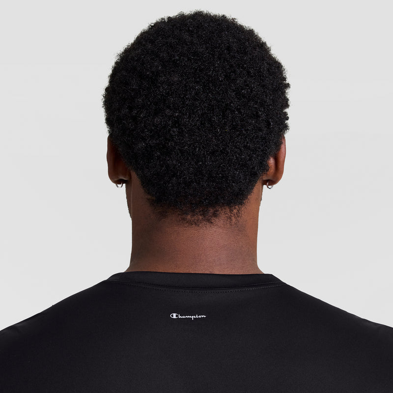 A person with short, curly black hair is seen from behind wearing a Champion Sport T-Shirt, Reflective C Logo - Big & Tall in black, featuring a small white reflective Champion logo on the upper back against a plain light background.