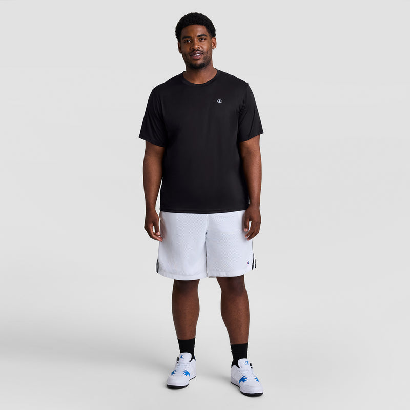 A man smiles against a light gray background, wearing a Champion Sport T-Shirt, Reflective C Logo - Big & Tall in black, white athletic shorts, black socks, and white sneakers with blue accents.