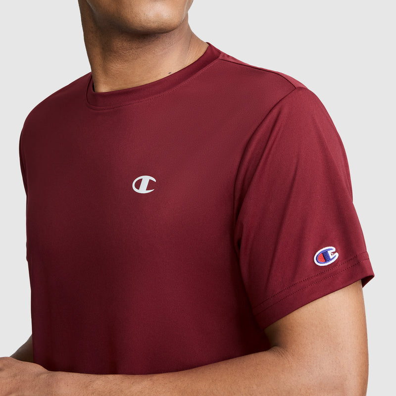A person is shown from the shoulders to mid-arm against a plain light background, wearing a maroon Champion Sport T-Shirt, Reflective C Logo. The men’s athletic shirt features the iconic reflective Champion logo on the chest and sleeve and is designed with moisture-wicking technology for lasting comfort.